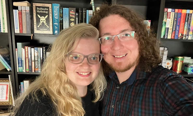 One the left side of the photo is Emily, a young woman with long blond hair, glasses and smile. On the right is Derek, her husband, a man with should length curly hair, glasses, and a smile. Behind them is a full bookcase.
